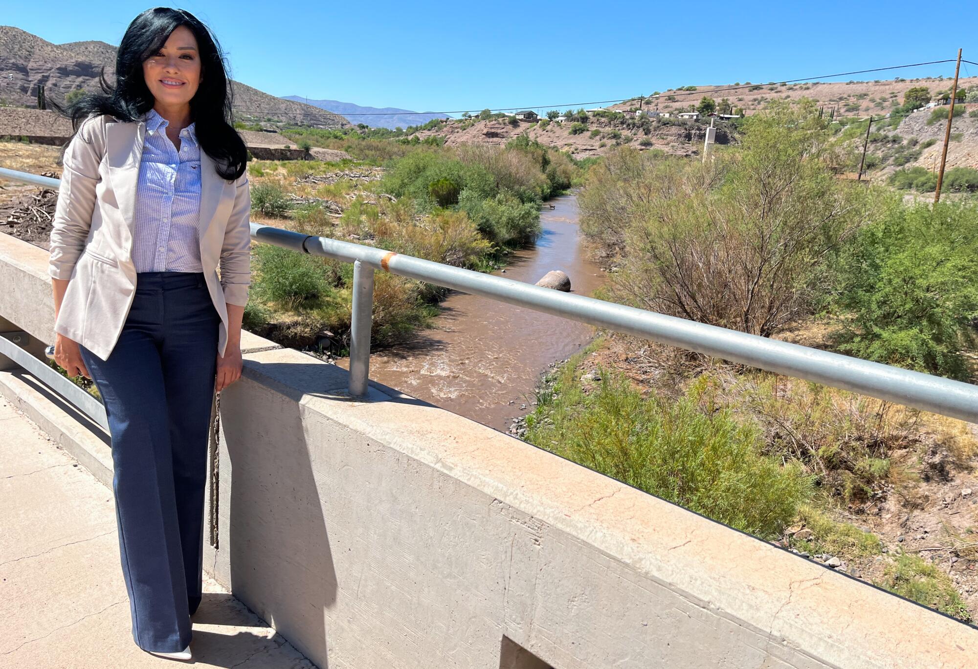 A woman with long black hair, in a cream jack and dark pants, stands on a bridge over a brown river in a desert landscape