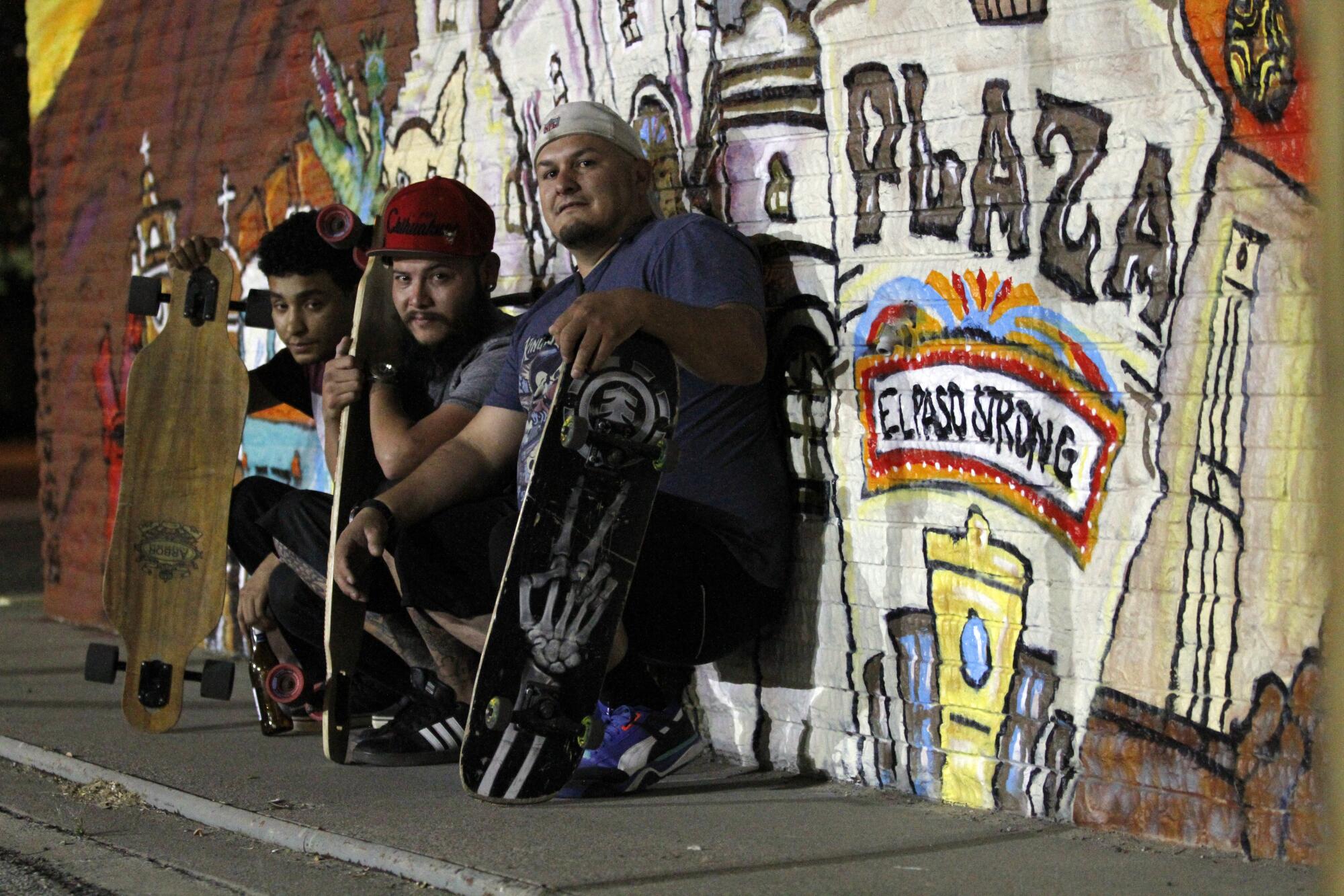 Three men with skateboards sit in front of a mural with the words "El Paso Strong"