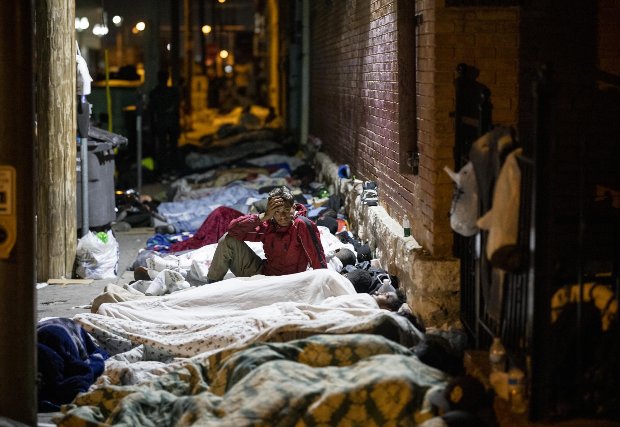 A man sits up, flanked by people sleeping in a row on a sidewalk next to a brick wall 