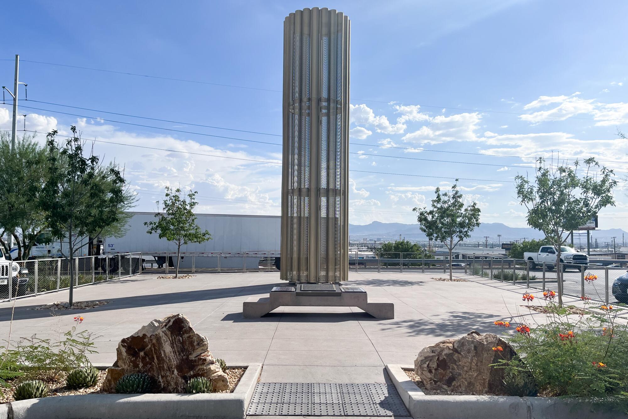 A column-like structure with a base stands at the center of a paved area, with some trees on the periphery