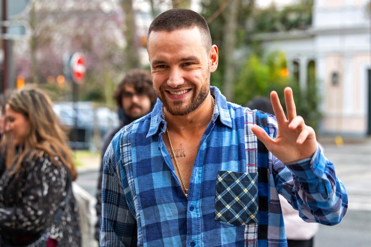 Liam Payne in a blue plaid flannel shirt smiling and waving with one hand