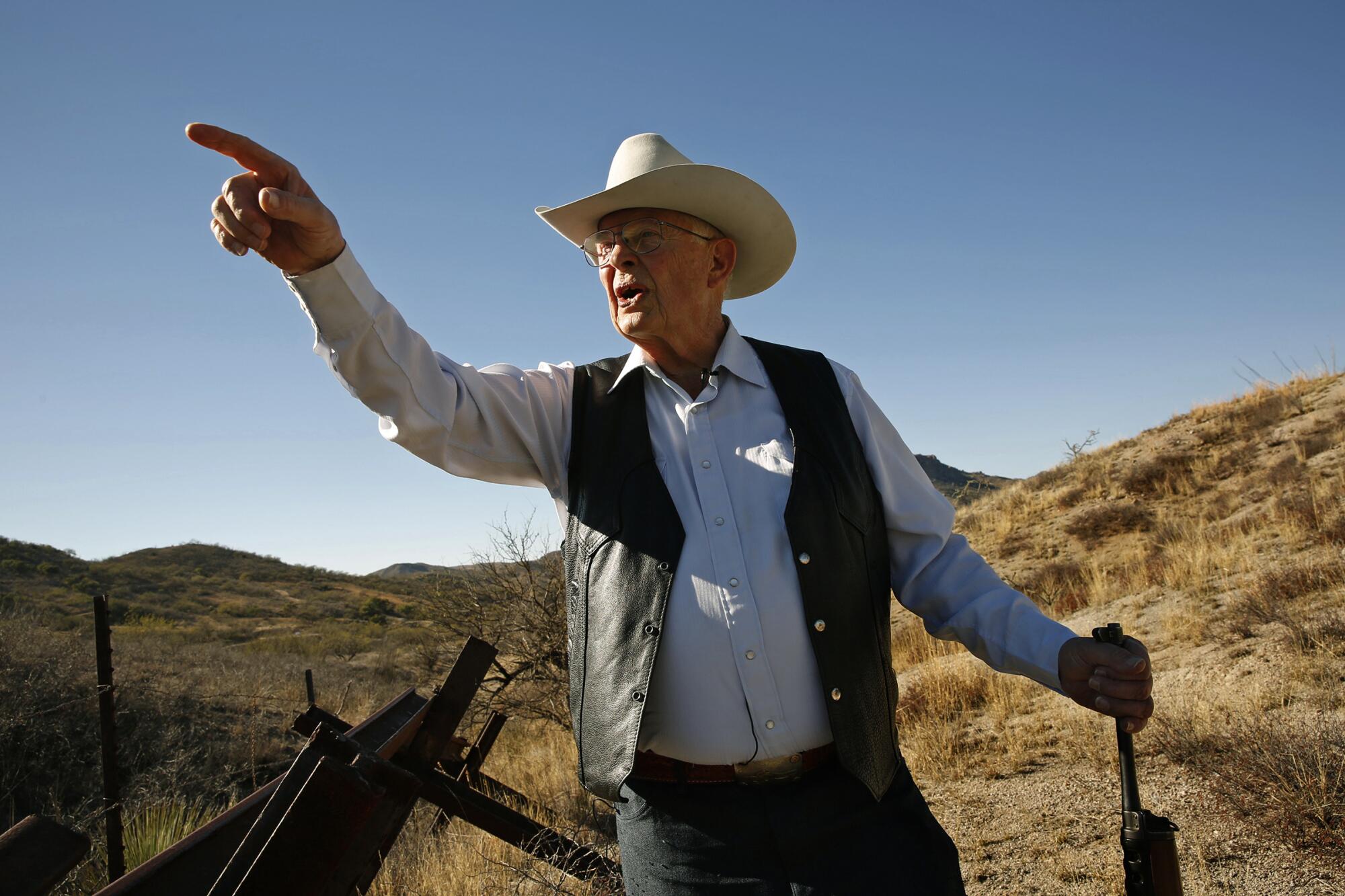 A man in a white cowboy hat, long-sleeve shirt and dark vest points while standing on desert terrain