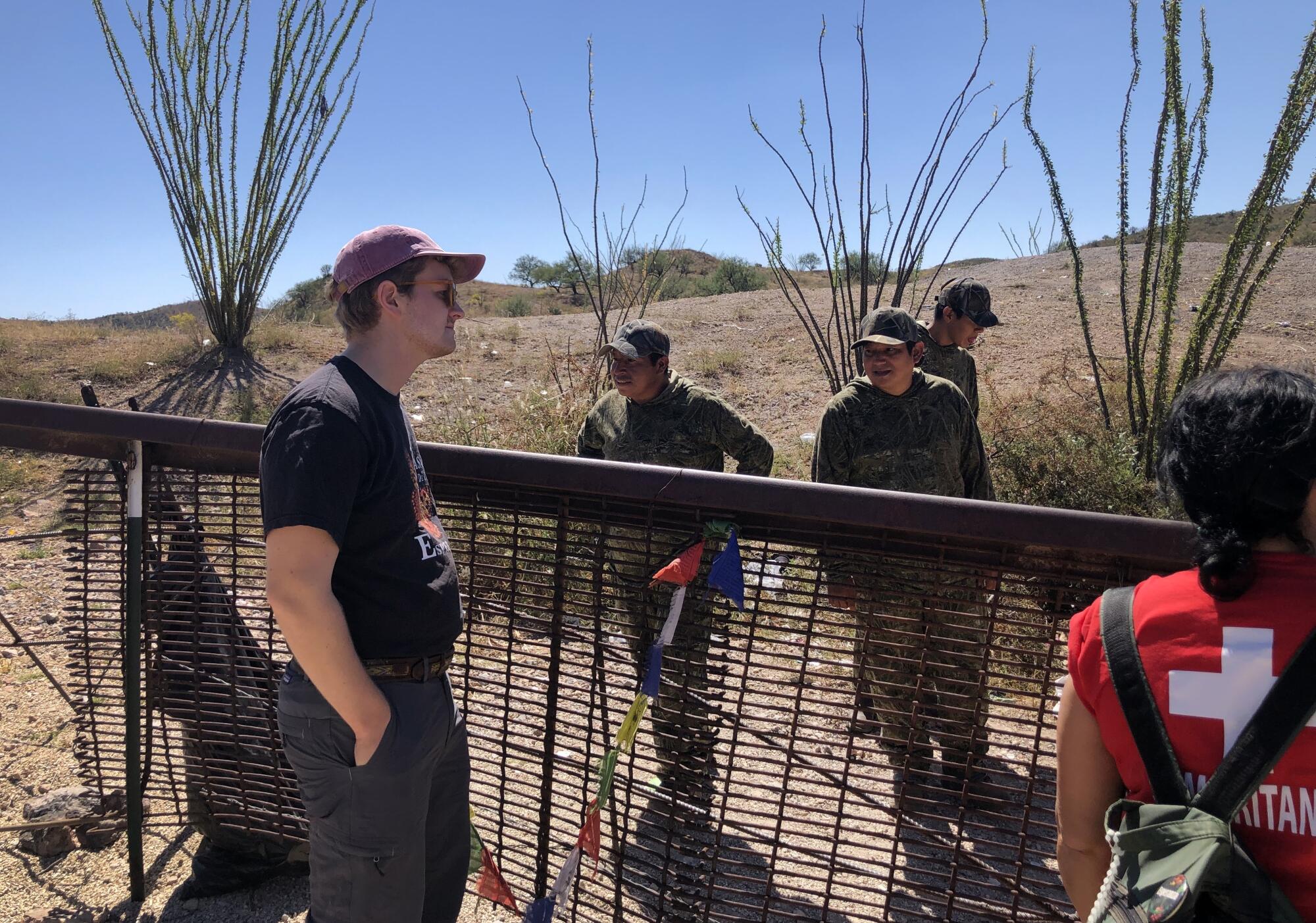 People stand on both sides of a wire fence in a desert setting dotted with bare, cane-like plants