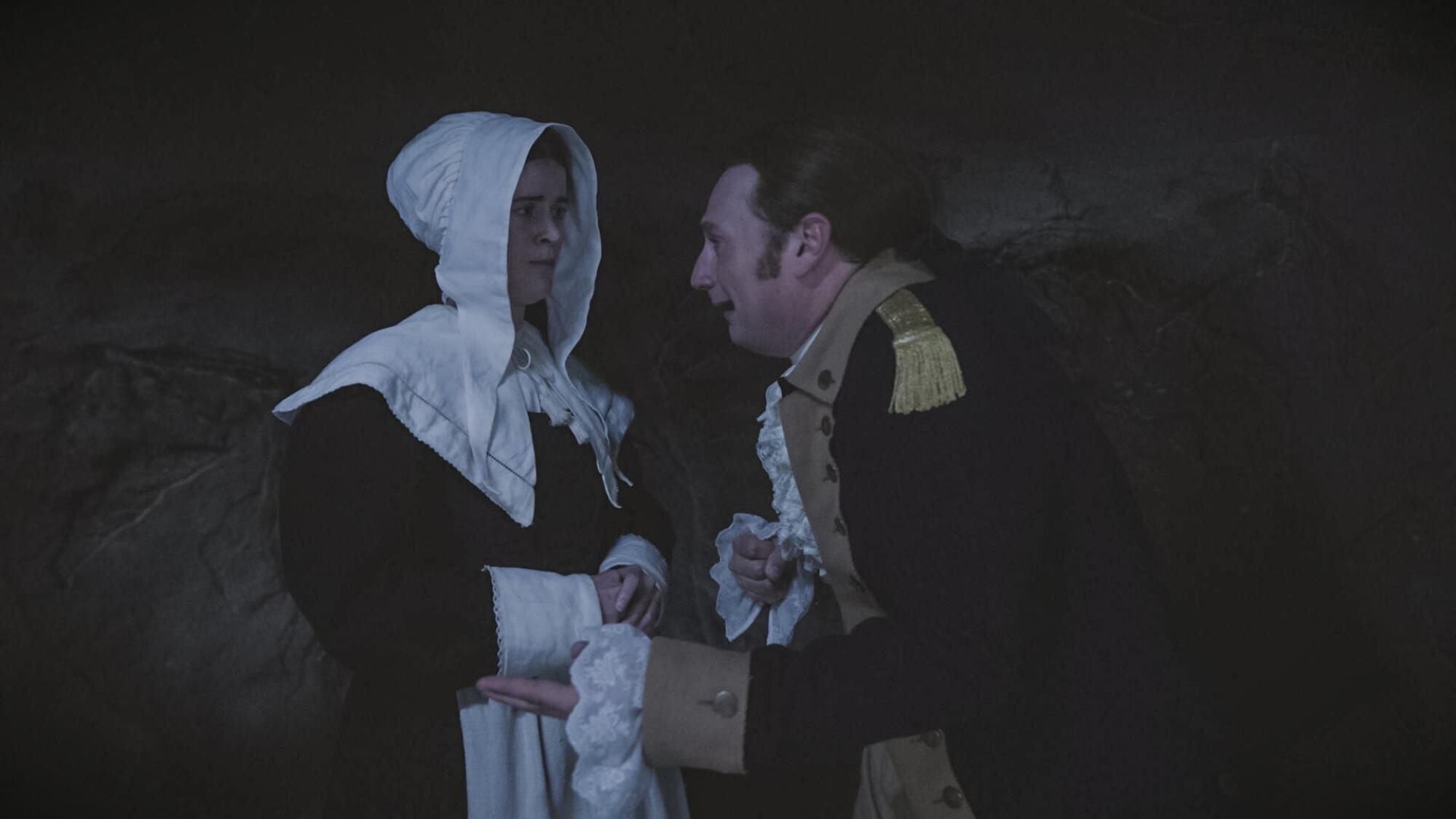 A woman in a white bonnet and dark dress looks at a man in a colonial military uniform.