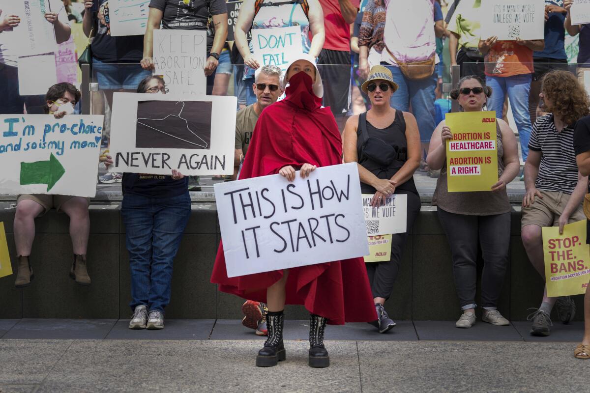 A person dressed in red as a character from "The Handmaid's Tale" holds a sign reading, "This is how it starts"