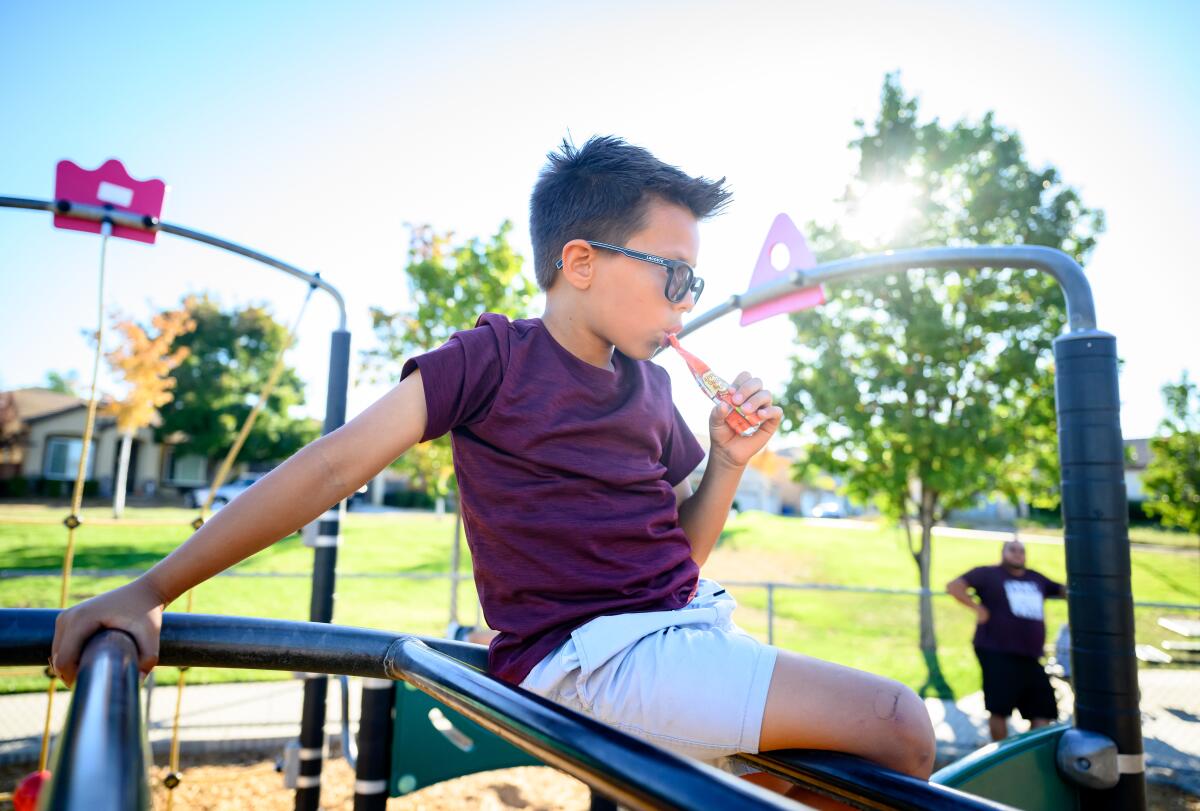 A boy eats baby food from a pouch while his father watches at a park