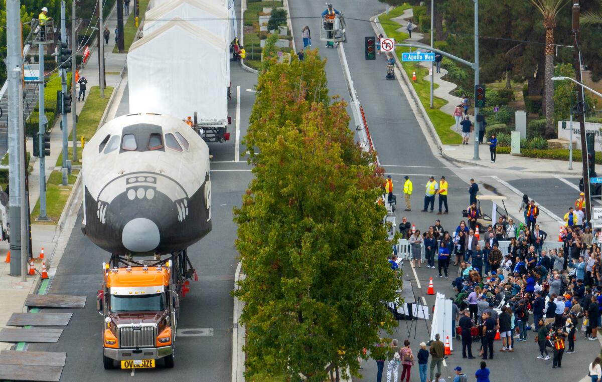 A to-scale model of a space shuttle moves along a road as people gather to watch.