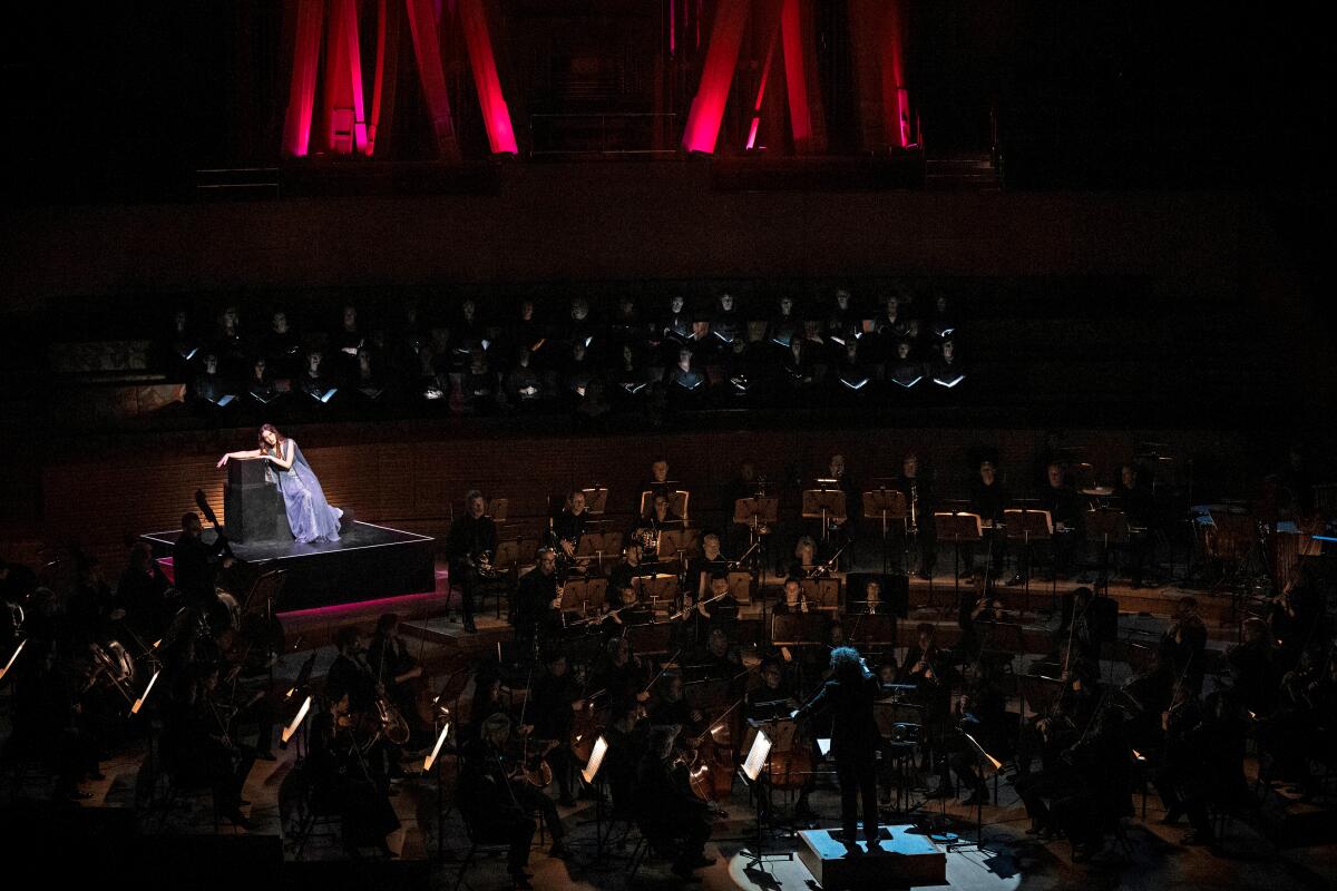 A woman illuminated on a platform set onstage among the members of the Los Angeles Philharmonic orchestra.