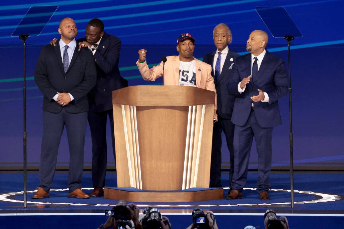 A man in a yellow jacket and dark cap raises a fisted hand while speaking at a lectern, flanked by four men in dark suits