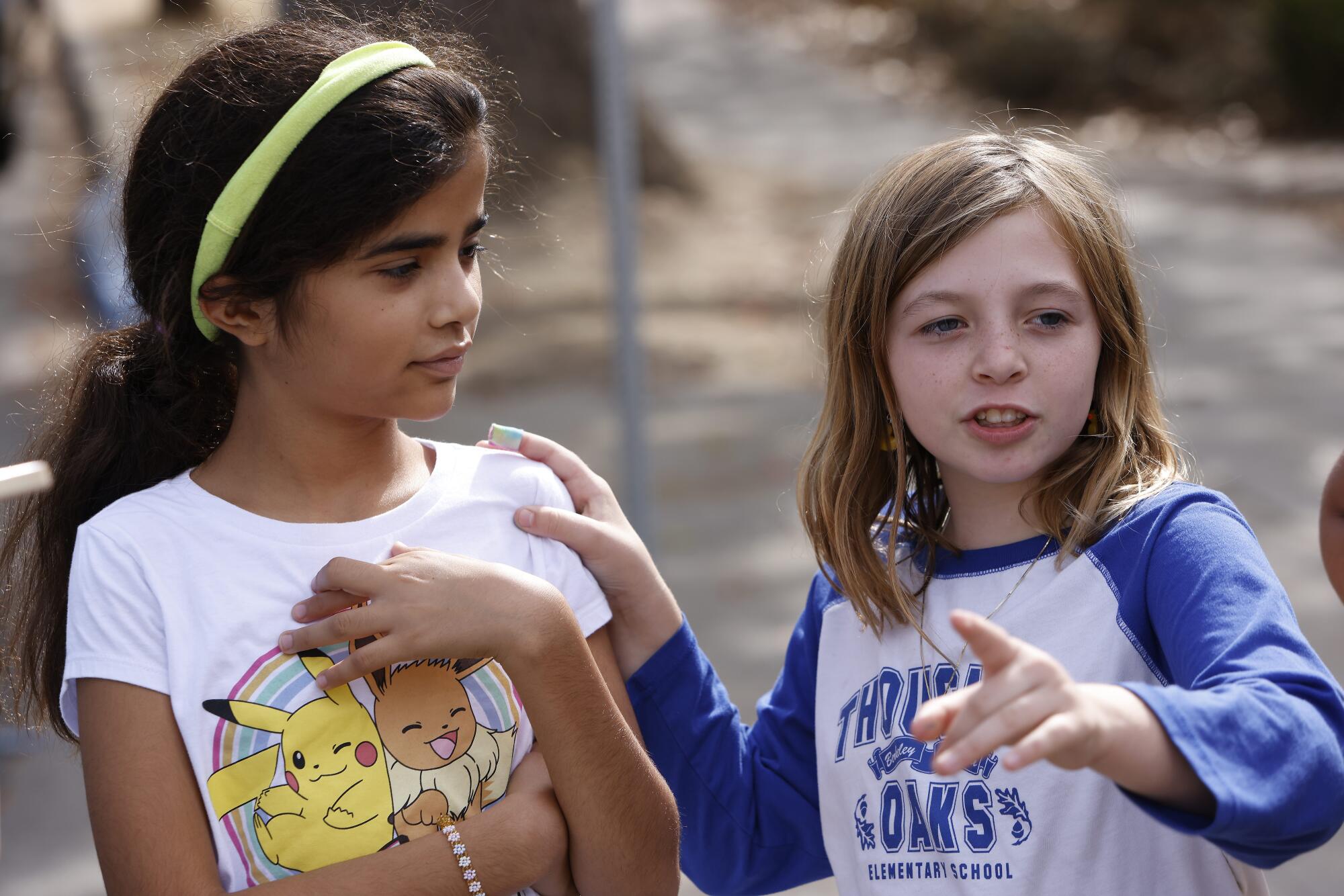 A young girl points as she speaks outdoors, one hand on the shoulder of another young girl listening to hear