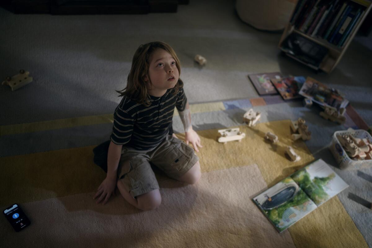 A little boy with shoulder-length hair sits looking up on a rug with toys around him.