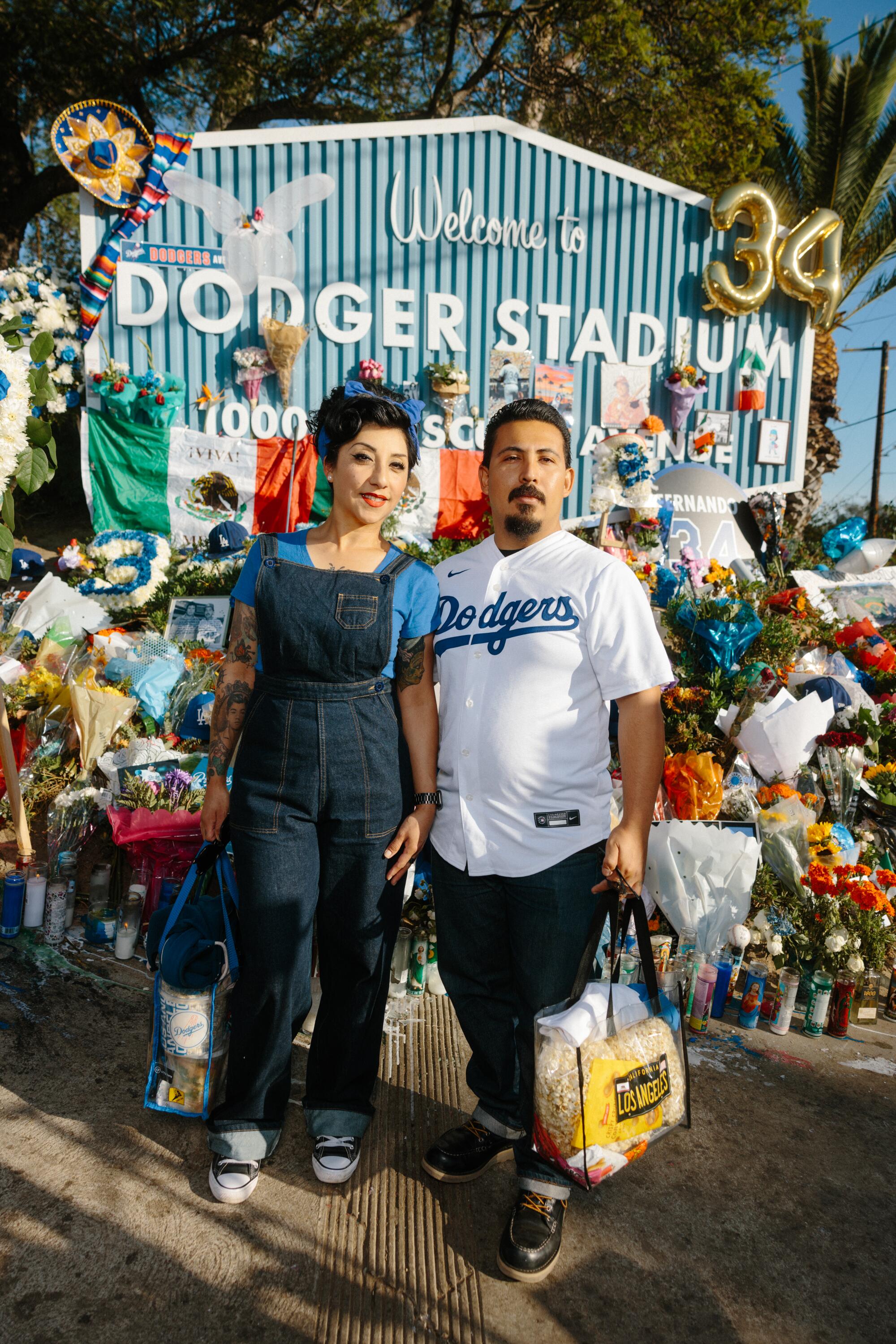 A man in a Dodgers jersey and a woman in overalls and a blue shirt stand in front of flowers at Dodger Stadium