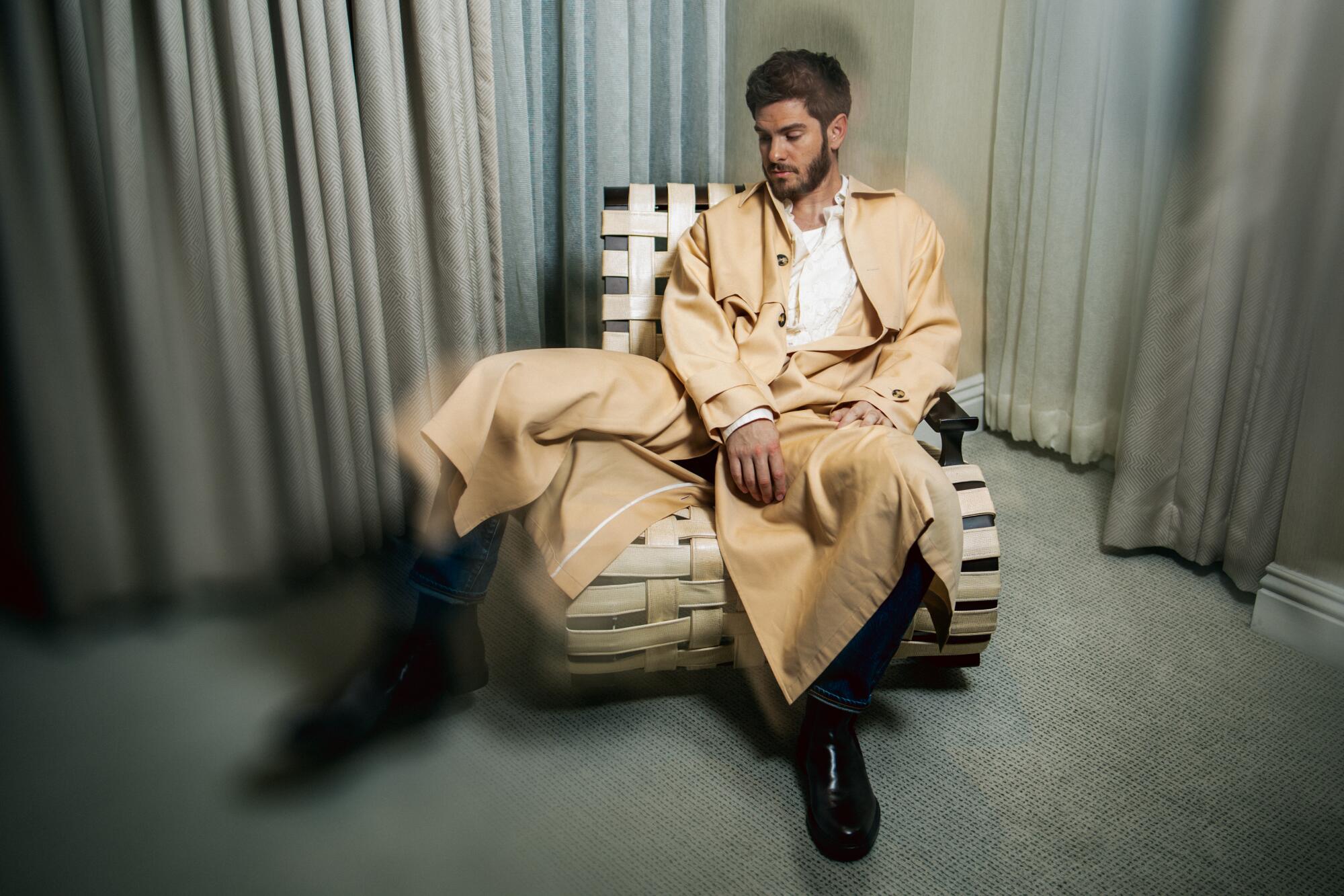 Andrew Garfield, wearing a beige suit with very wide-legged pants, sits on a curving woven chair