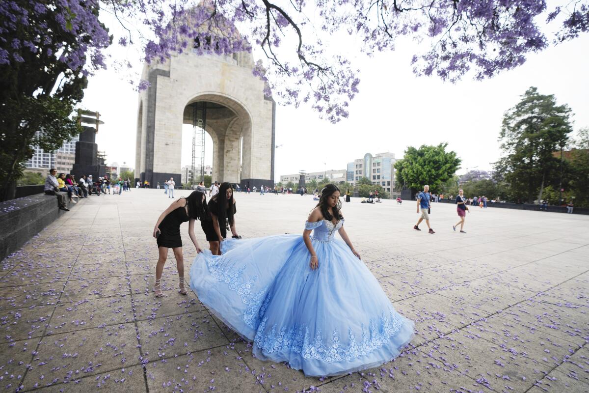 Two people hold up the train of a light blue gown worn by a girl near a monument that forms an arch