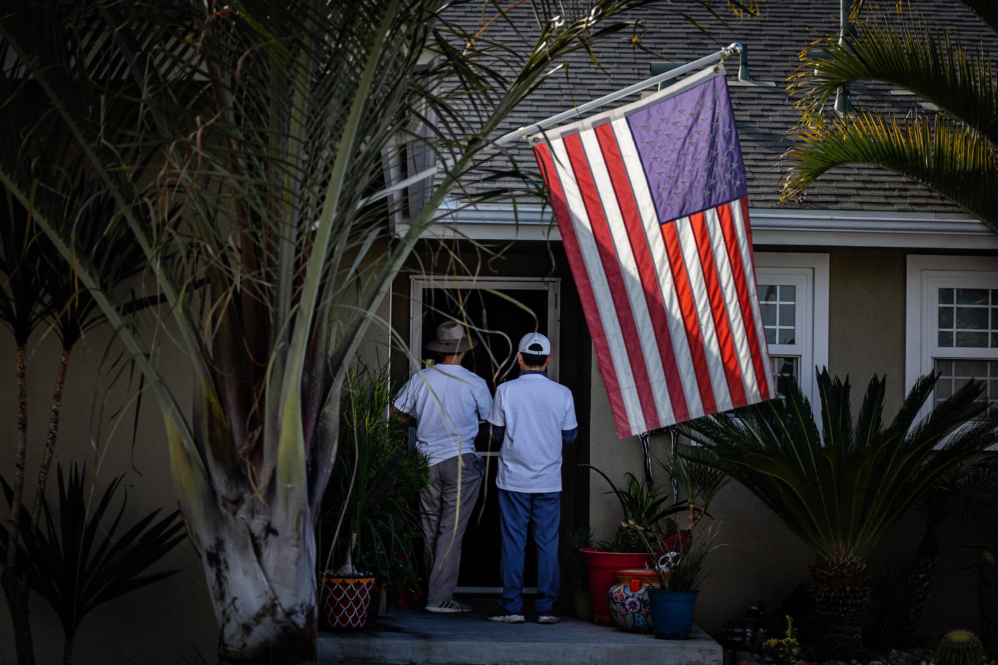 Two people stand at a home's entrance under a U.S. flag.