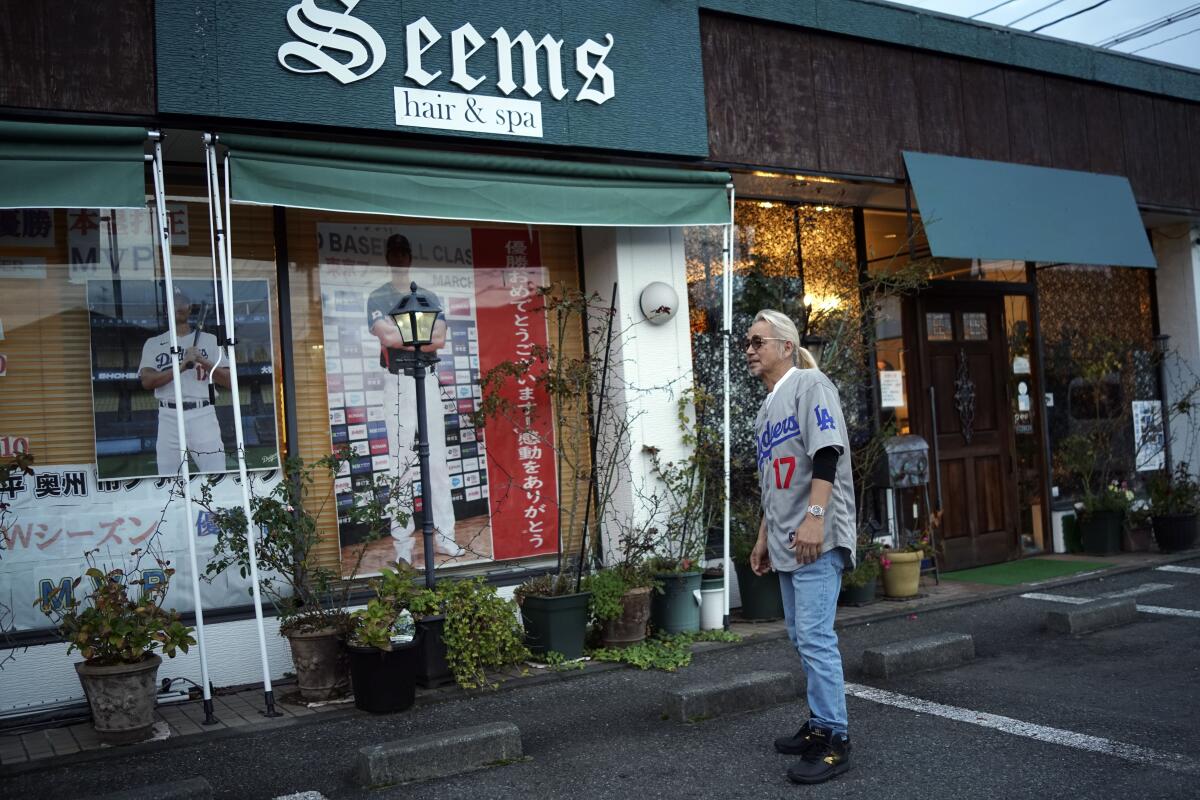 A man stands outside building whose front display shows baseball memorabilia. Its sign reads "Seems hair & spa." 