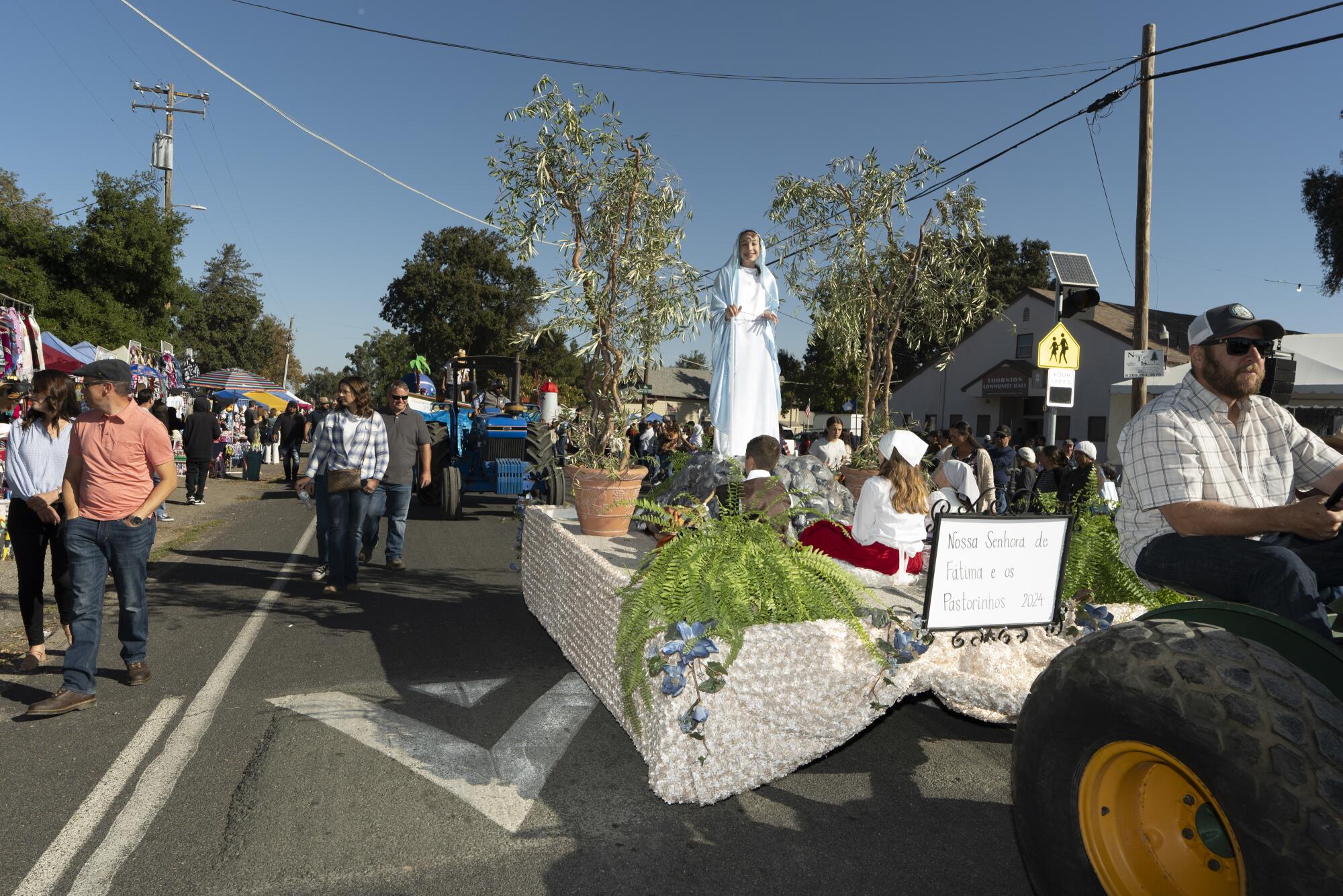 A girl in a white dress stands on a platform pulled by a tractor during a parade