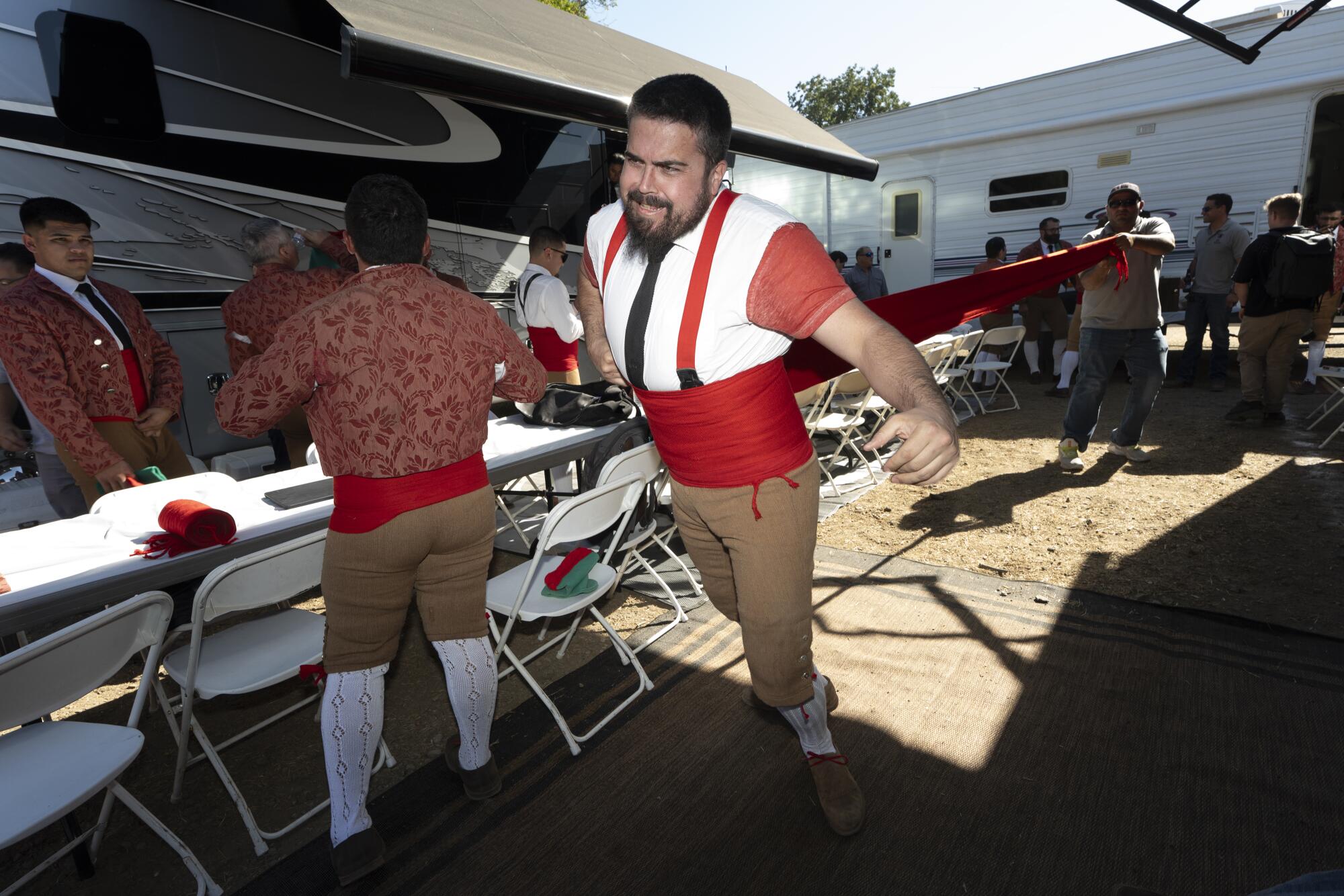 A man with a dark beard, wearing a red cummerbund and khaki pants, stands near other men at a long table with chairs