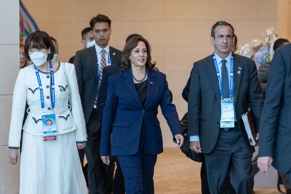 A woman in a dark blue pantsuit is flanked by a masked woman in a white outfit, left, and a man in a dark suit and tie