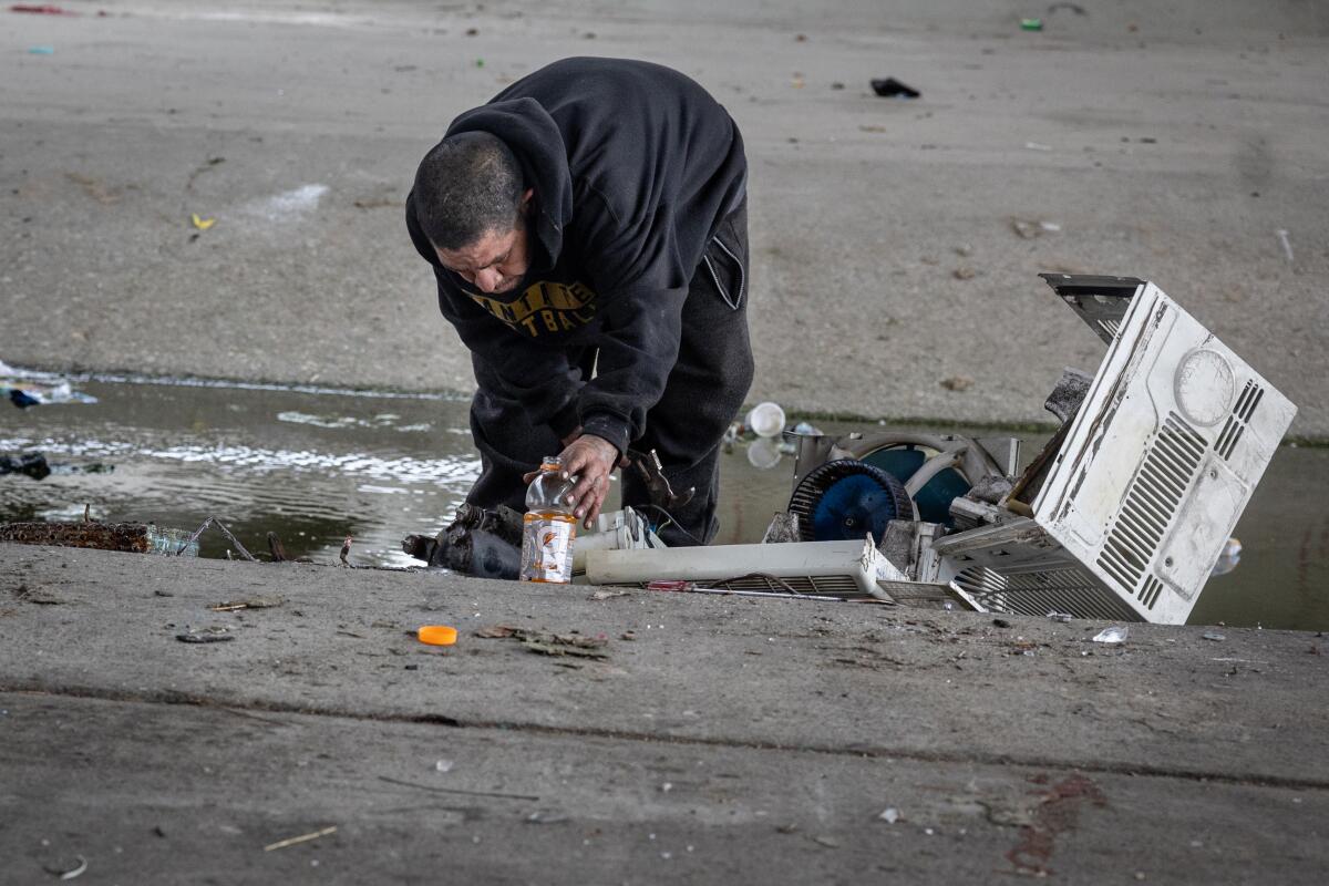 A man reaches toward a sports drink bottle in a trash strewn concrete channel.