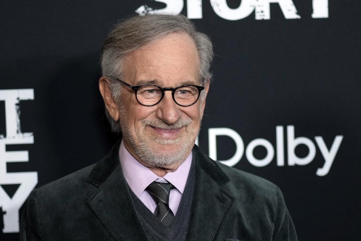 A man with black-framed glasses and gray hair smiles for cameras at a movie premiere