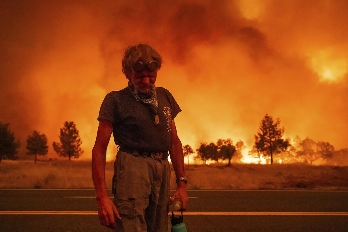 A person in a dark T-shirt and khaki pants looks down as smoke and orange flames billow behind him, with trees in silhouette