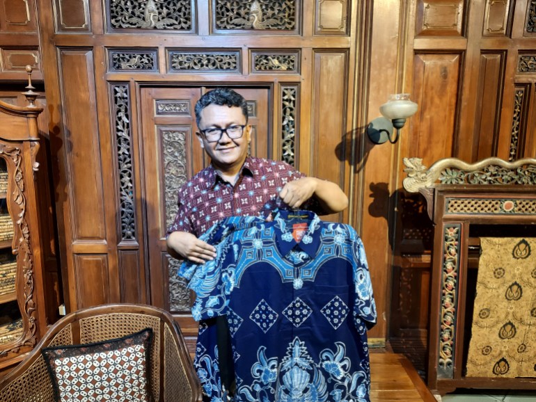 Gunawan Setiawan. He is wearing a marron-coloured batik short and holding up shirts with blue designs, He is in a wood-pannelled room with rattan-sated chairs. 