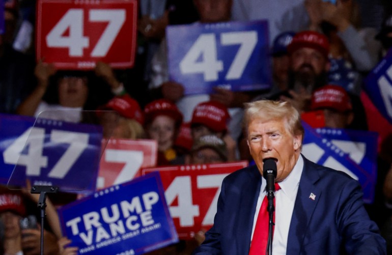 Donald Trump speaks at a rally, as people hold up signs that read "47" behind him.