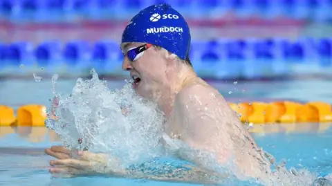 PA Media Team Scotland athlete Ross Murdoch mid breast stroke during Glasgow 2014. He is wearing a dark blue swimming cap and goggles.