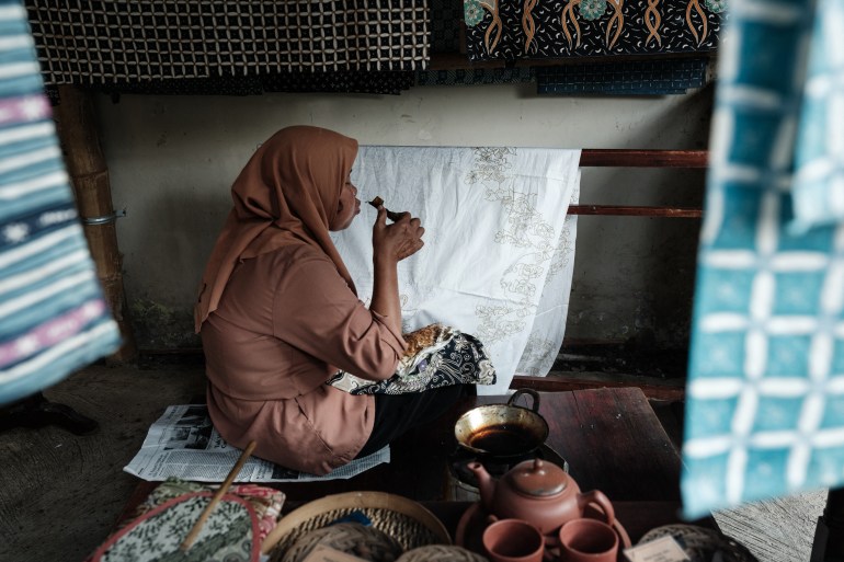 A woman draws wax pattersn onto a while cloth with a traditional tool. The cloth is hanging from a pole