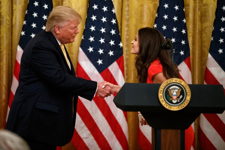 Donald Trump shakes hands in the White House with Lila Rose, who stands behind a podium with the presidential seal. Behind them is a row of US flags.