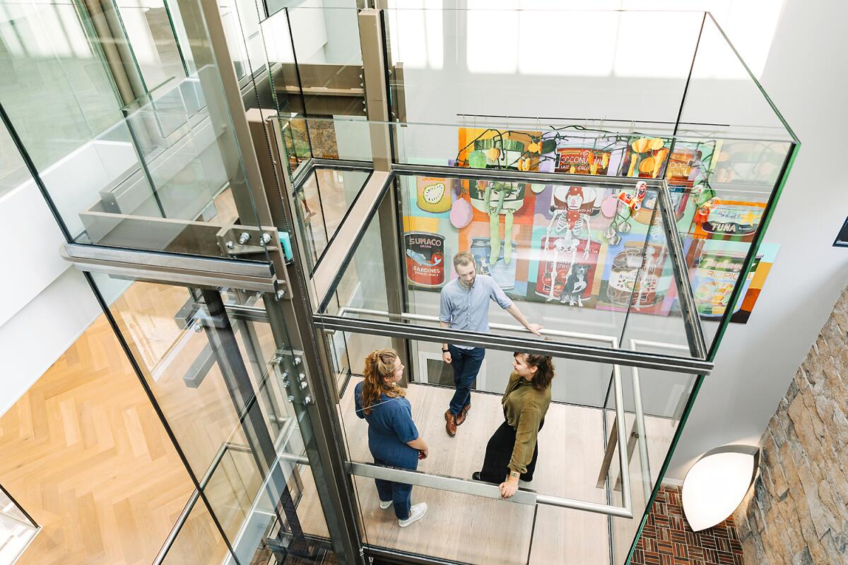 Three people are seen from above riding in a glass elevator in the middle of a room at an art museum