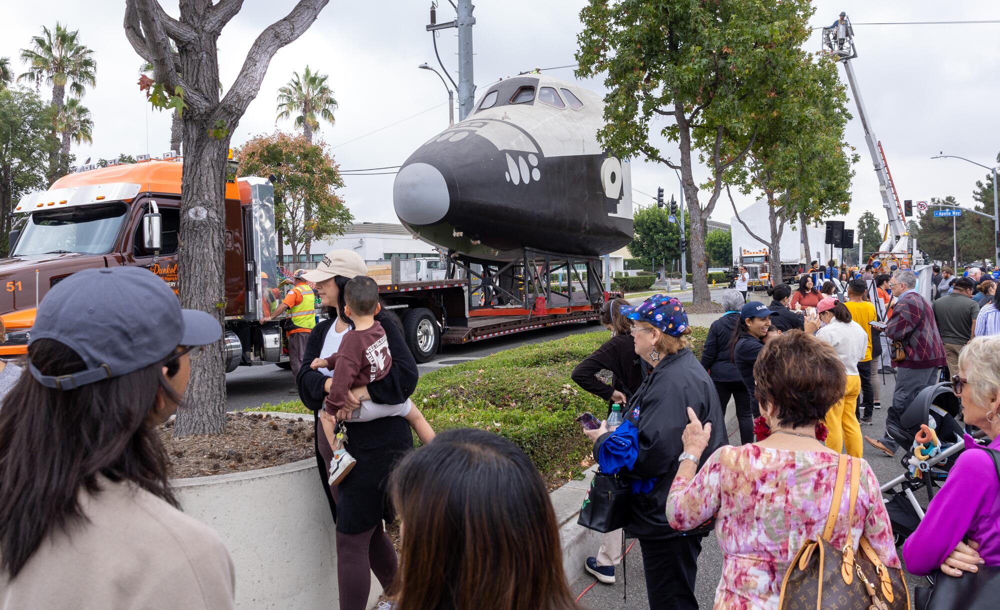 People watch as a truck carrying a space shuttle model passes on the other side of a divided road.