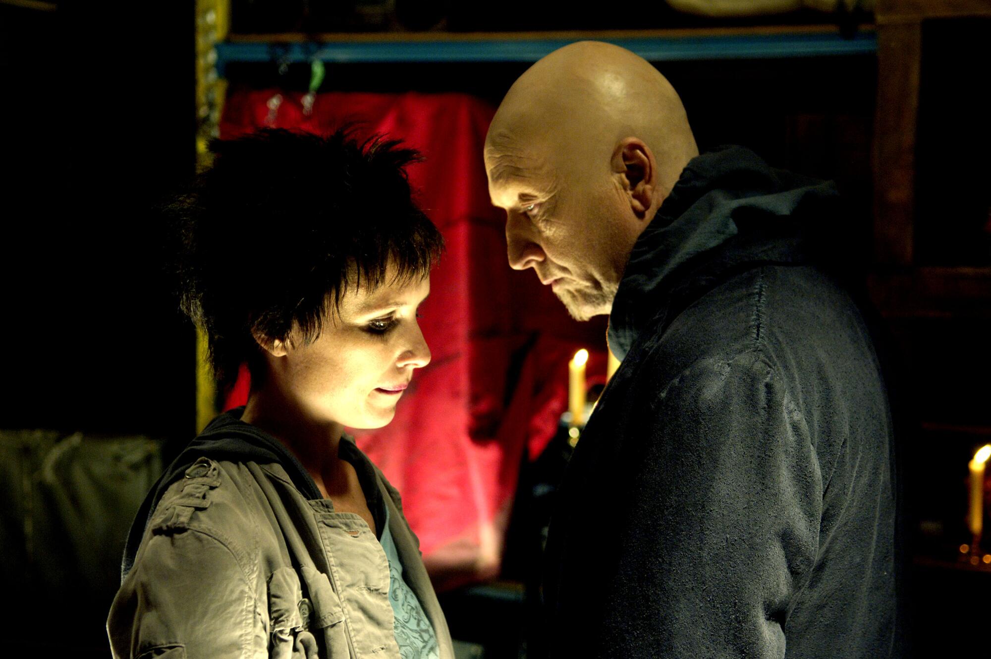 A bald-headed man speaks to a dark-haired woman.