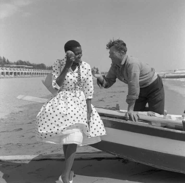 South african singer and civil rights activist Miriam Makeba, wearing a polka-dotted dress and holding a handbag, portrayed while sitting on a boat and listening to the sea sound in a shell, a man smiling to her, Lido beach, Venice 1959 (Photo by Archivio Cameraphoto Epoche/Getty Images)