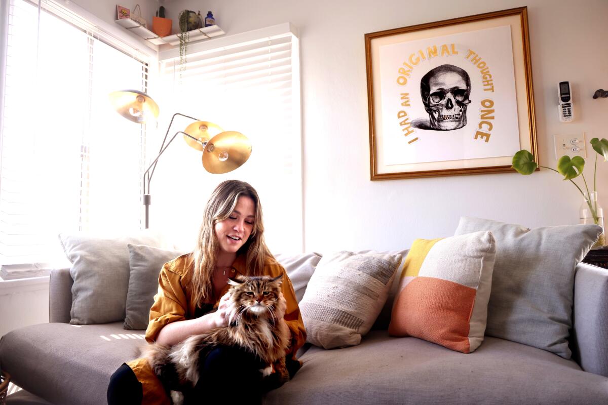 Ariel Kaplan sits on a gray couch and pets her fluffy cat.