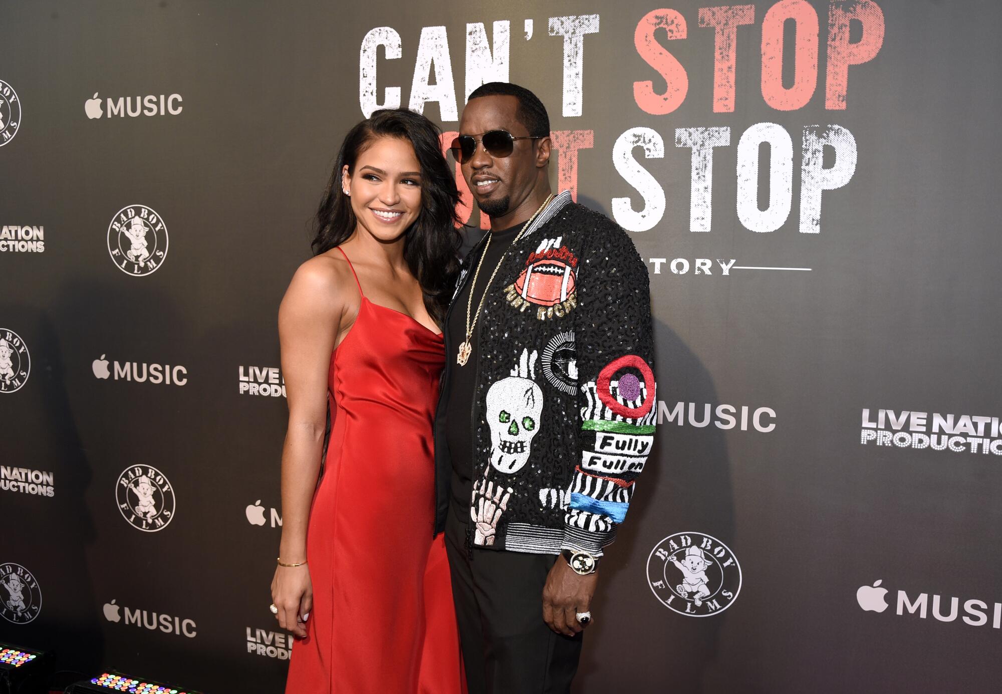 Cassie in a red sleeveless gown posing next to Sean "Diddy" Combs in a black jacket and sunglasses at a red-carpet event