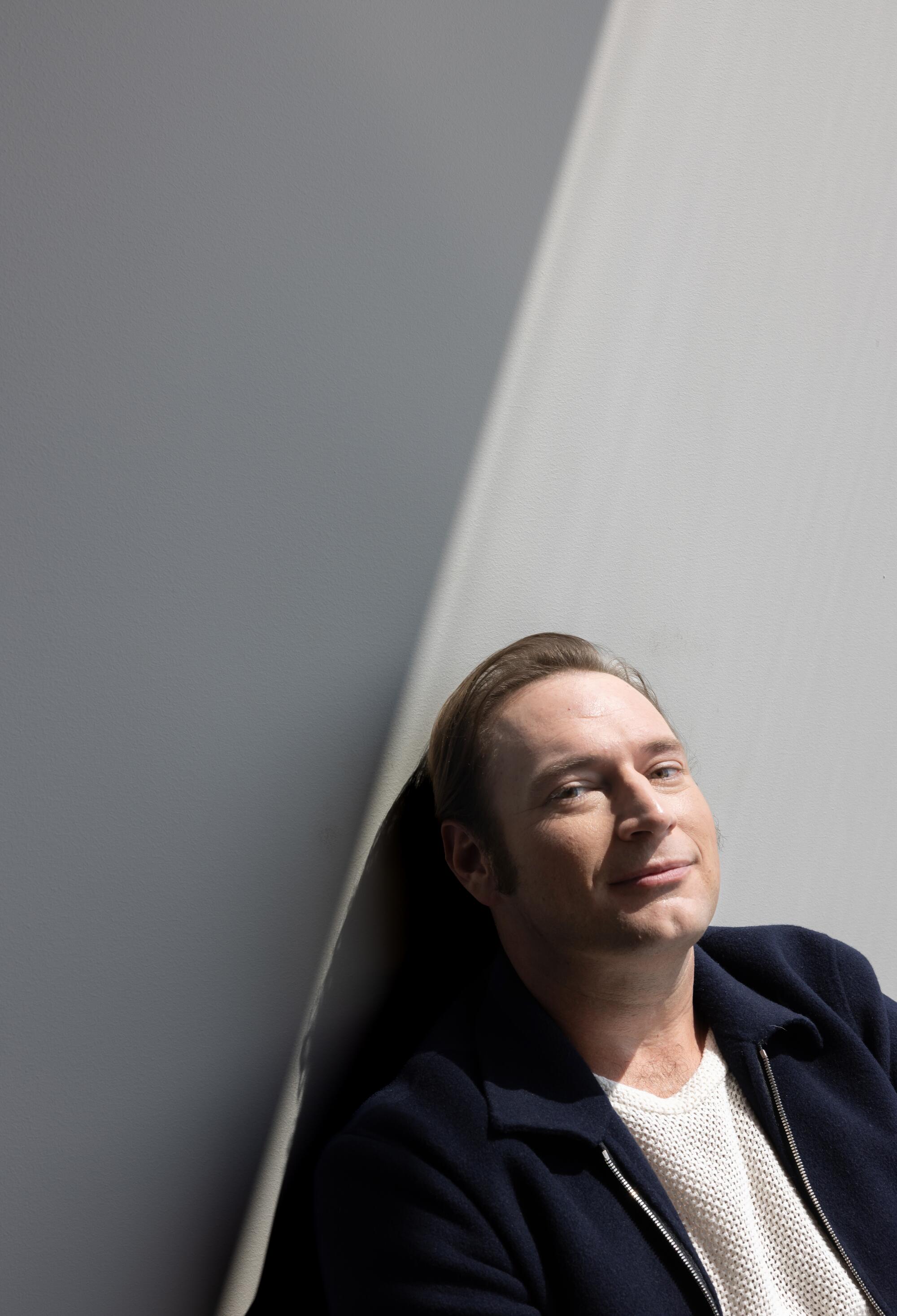 A man in a white T-shirt and dark jacket leans against a wall with a shadow cast across it.