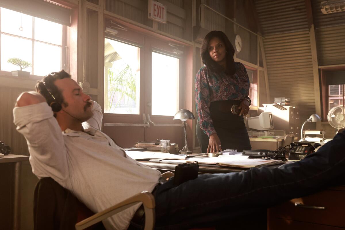 A man leans back in his desk chair listening to headphones while a woman stands and watches him.