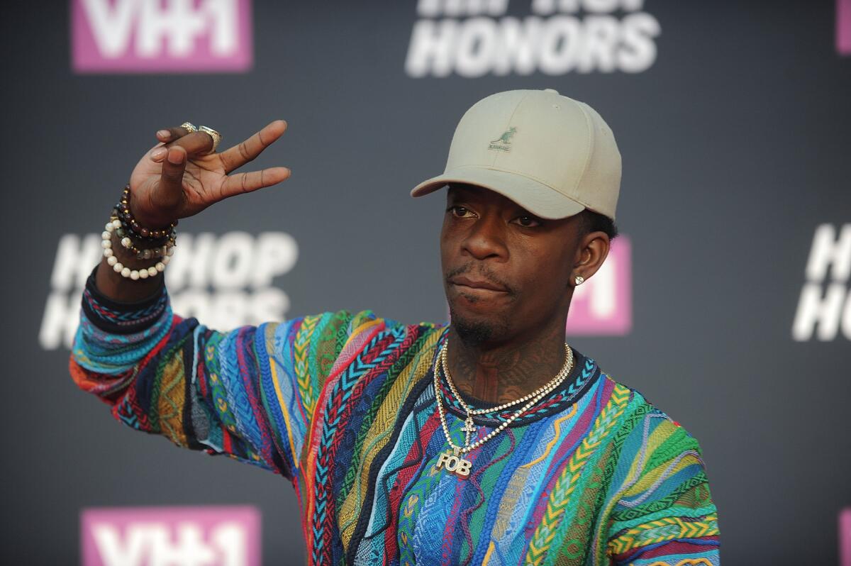 Rich Homie Quan making a peace sign with one hand while clad in a multicolor sweater and beige baseball cap