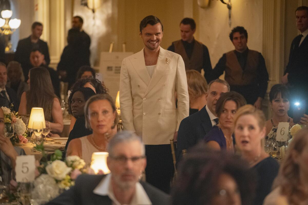 A man in white dinner jacket over a white T-shirt stands among a seated crowd