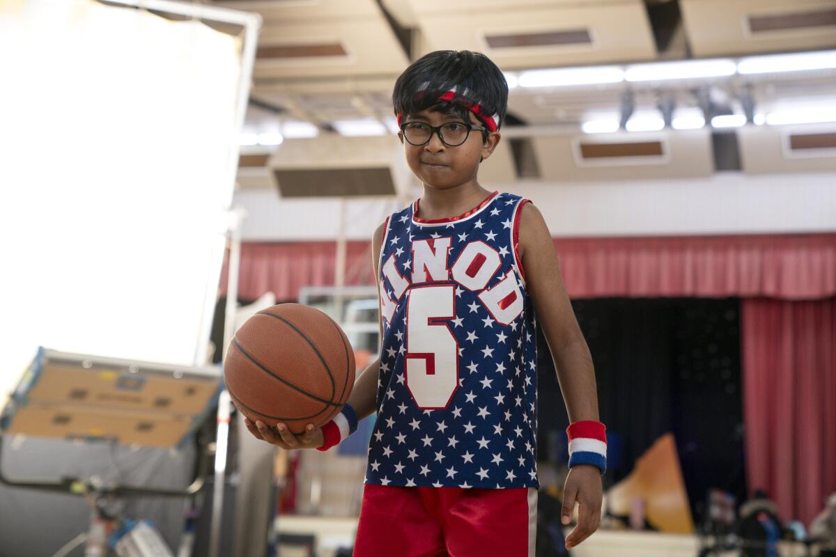 A boy with glasses in a red, white and blue basketball uniform holds a basketball in one hand.