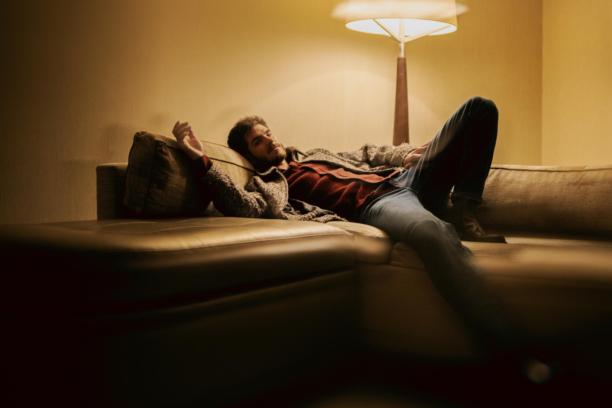 Andrew Garfield sprawls on a couch under the light of a table lamp