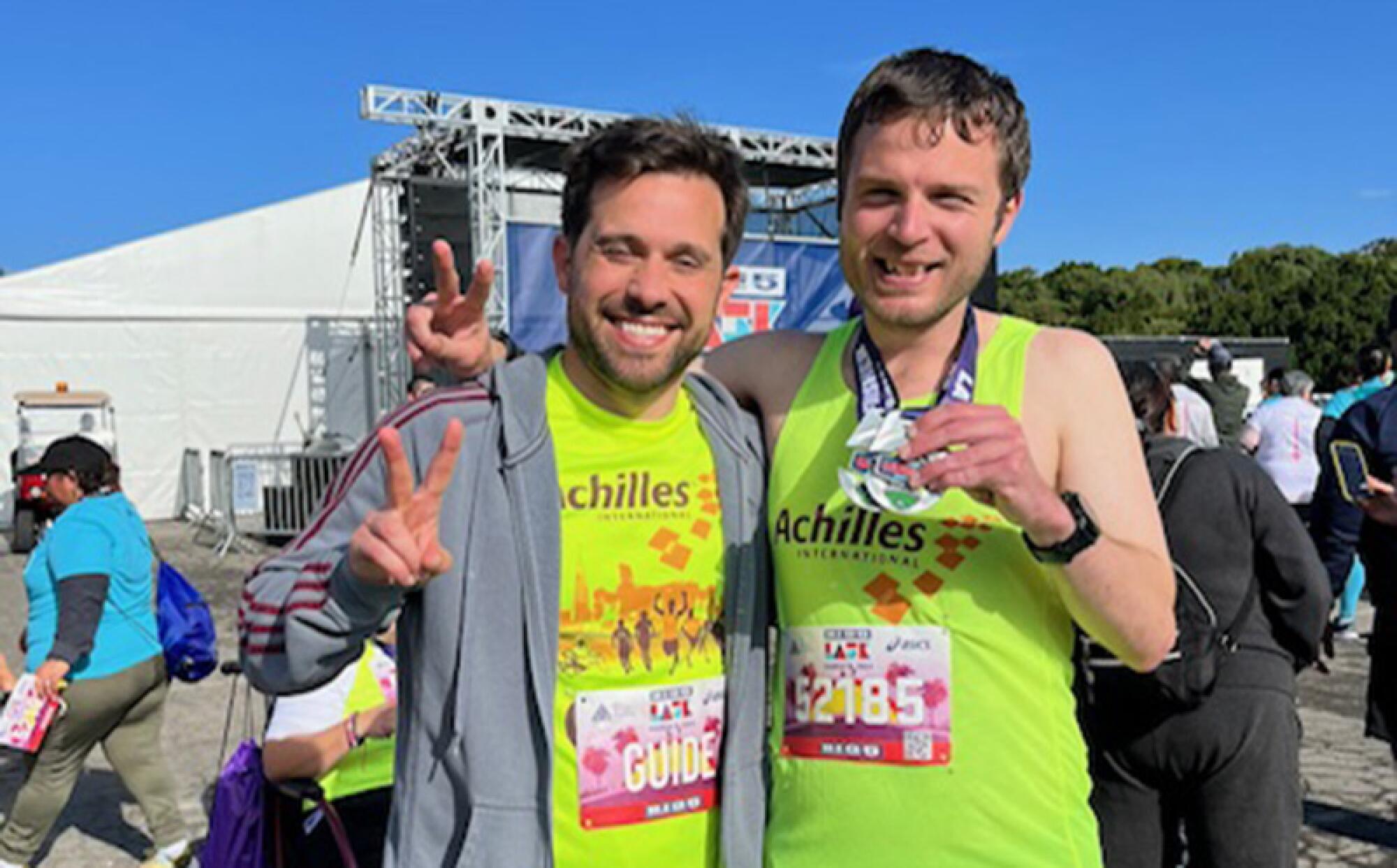 Two people outdoors in neon green Achilles shirts flash peace signs. One has medals around his neck.