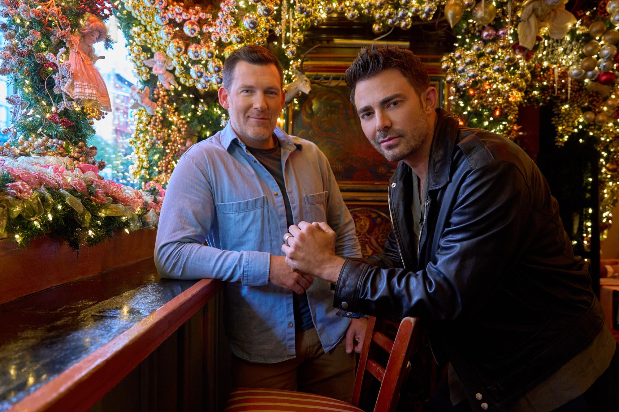 A man in a denim shirt leans on a bar and a man in a black leather jacket stands next to him.