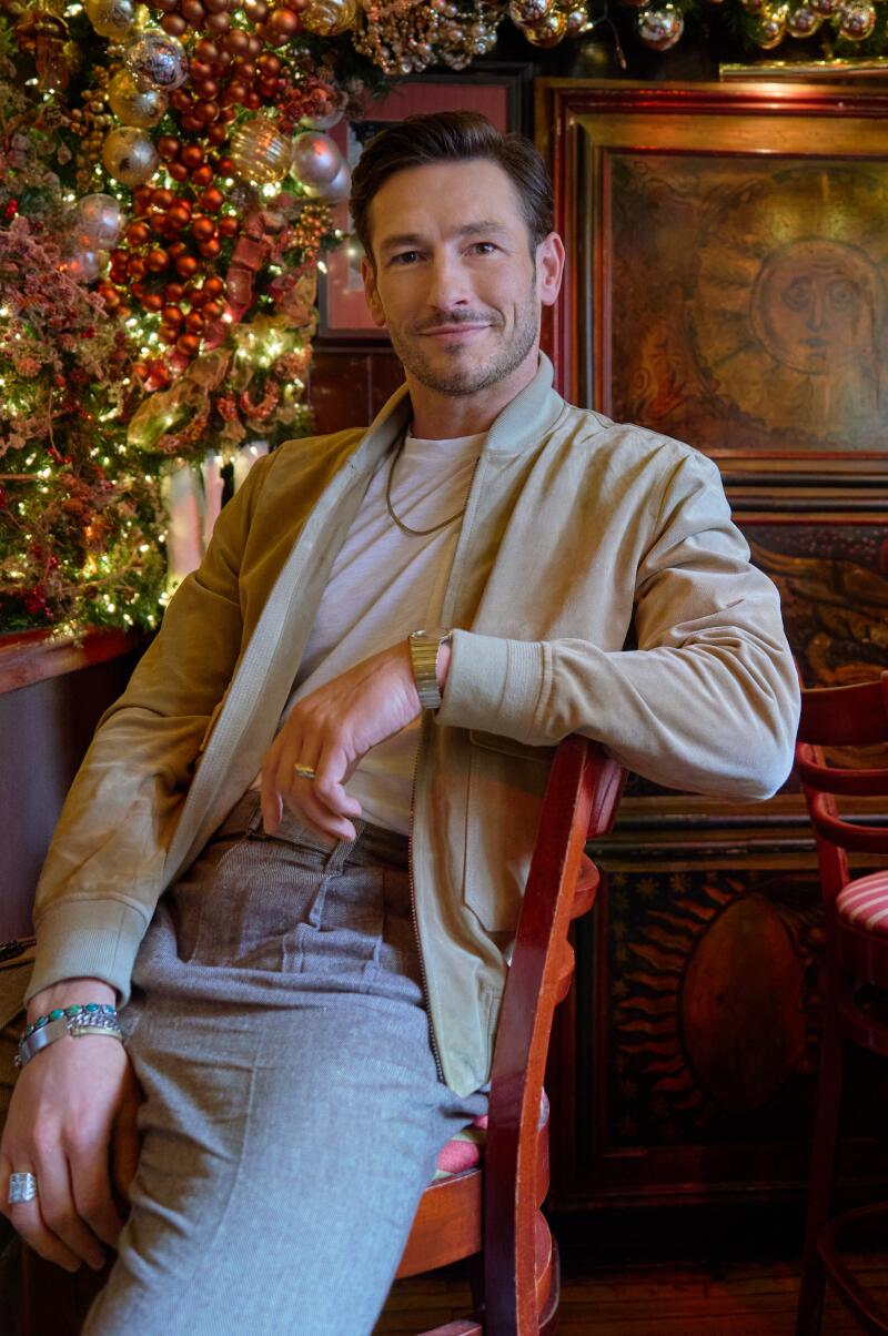 A man in a tan jacket, white T-shirt and gray pants sits on a bar stool with an arm leaned on the back.