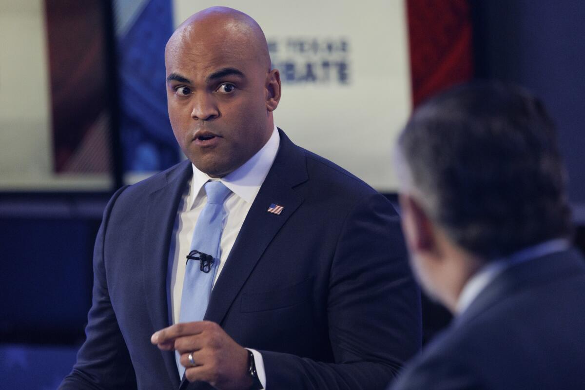 A man with a clean-shaven head, in dark suit and light blue tie, speaks while looking at a man, seen from behind 