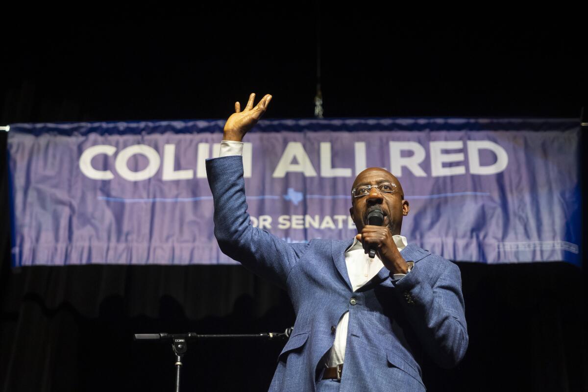 A man with glasses, in a blue-gray jacket and white shirt, raises one hand while speaking into the mic in his other hand