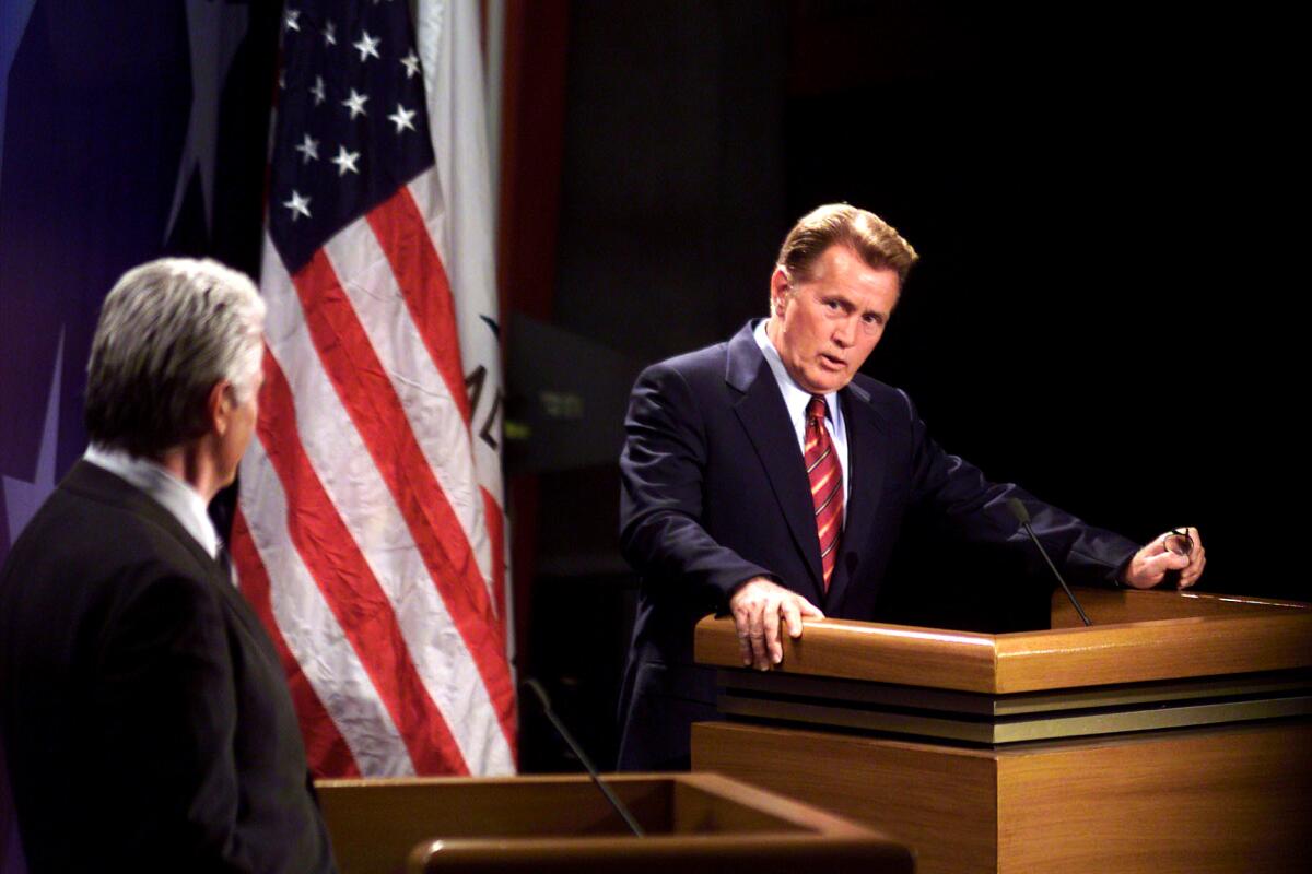 Two men stand at lecterns in a scene from "The West Wing."