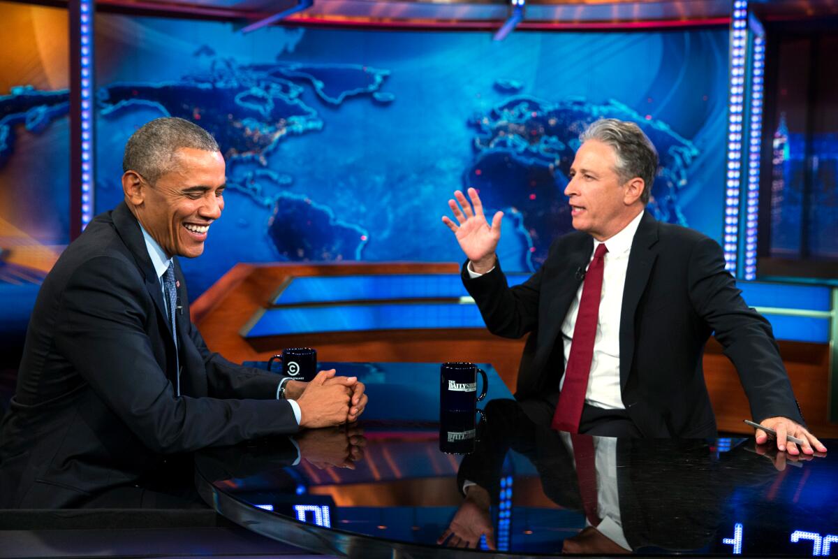 President Obama sits at a table with Jon Stewart with a world map in the background during a "The Daily Show" taping.